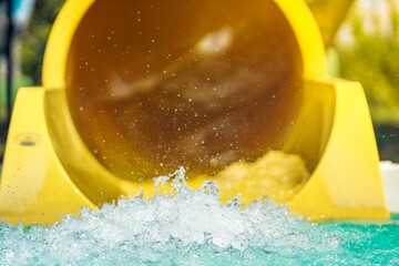 Families enjoy a bright summer day at a vibrant water park. Children and adults laugh as they zoom down a yellow slide, causing splashes in the inviting pool below