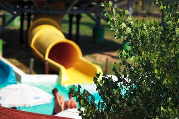 Families visit a lively water park on a warm afternoon. Laughter fills the air as children slide down the bright yellow water slide while parents relax under umbrellas nearby