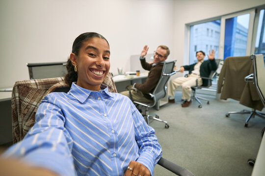 Portrait of young adult Hispanic woman smiling and taking selfie in modern office while two Caucasian men, one middle aged and one young adult, sitting at desks waving in background