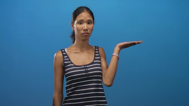 Thai woman with palm up presenting gesture and bare hand visible, wearing a striped tank top in a blue studio; uncertainty contemplation.