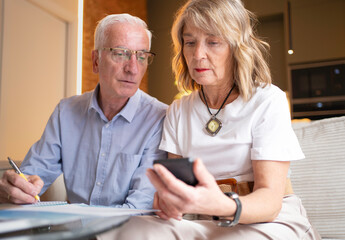 Senior couple analyzing finances and budget using a smartphone and writing notes at home
