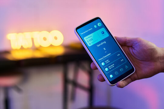 Caucasian young adult man holding smartphone displaying digital banking app interface with transaction details visible, blurred tattoo sign in background, right hand in focus