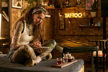 Young adult Caucasian woman with extensive tattoos sitting cross legged on table drawing in...