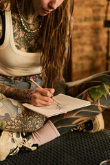Young adult Caucasian woman with extensive tattoos sitting cross legged sketching in notebook,...