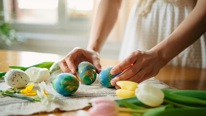 Woman's hands arranging hand painted easter eggs with intricate designs and vivid colors on a table with tulips and daffodils, creating a warm, festive spring display