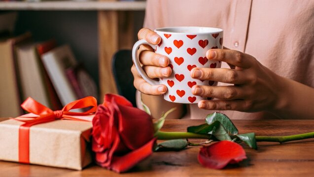 Woman holding heart pattern mug, enjoying hot drink with a red rose and a wrapped gift nearby, celebrating romance, love, and special occasions like valentine's day - Powered by Adobe