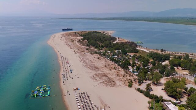 Aerial drone pan capturing the white sand and turquoise waters of Keramoti Blue Flag Beach, Kavala, Greece, with coastal pine forests and a distant ferry crossing.