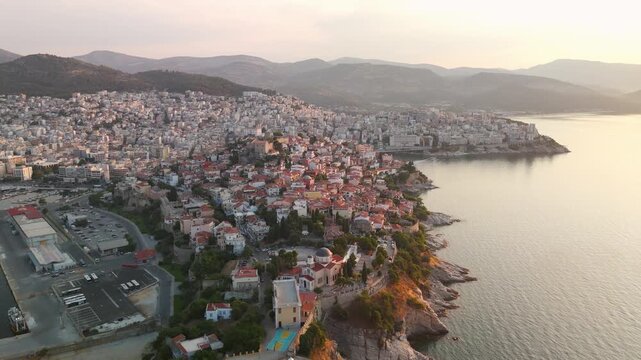 Spectacular high altitude aerial pan over the sprawling coastal city of Kavala, revealing the historic Panagia Peninsula, Acropolis Fortress, and Thracian Sea at golden hour sunrise.