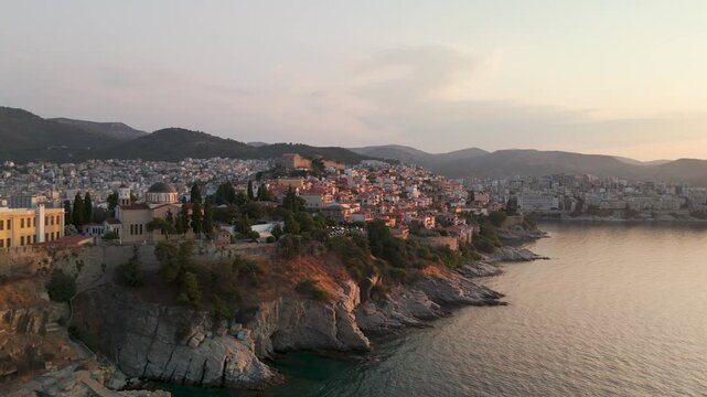 Aerial pan at sunrise over the historic Panagia Peninsula and Old Town of Kavala, Greece, highlighting the colorful houses and the Acropolis Fortress at the peak.