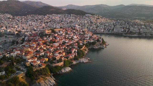 High altitude aerial drone pan revealing the expansive city of Kavala and the historic Panagia Old Town peninsula bathed in golden sunrise light, Eastern Macedonia, Greece.