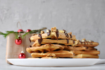 A tall stack of golden waffles topped with bananas, walnuts, and chocolate drizzle sits on a white plate, with festive Christmas decor and red ornaments softly blurred in the background.