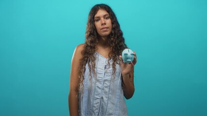 Teen woman holding blue piggy bank in her hand and showing it to camera in a studio setting with turquoise backdrop; financial contemplation.