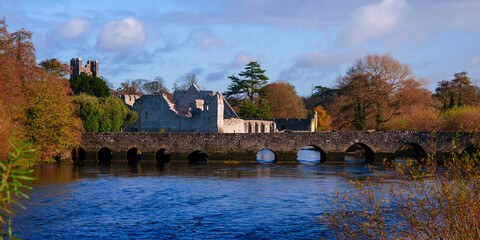 The Desmond Castle and the landmark old stone bridge, built in 1390, spanning the River Maigue in Adare, Ireland, in a vibrant autumn landscape.