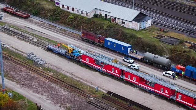 Aerial view of Poprad-Tatry rail yard: red maintenance train with cranes loading steel beams, blue service wagon, tanker car, workers in hi-vis, parked cars, and sidings with construction materials.