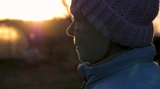 Profile view silhouetted elderly woman breathing out steam into cold air. Sunset with golden light and lens flare provides warm contemplative background. Warm golden light outlines rural female figure