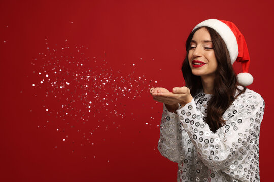Beautiful woman wearing Santa hat blowing confetti on red background. Christmas party