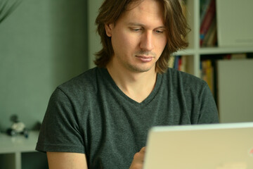 A young man with long hairstyle working on his laptop at home office desk. Focused look at the computer screen. Remote work and telecommuting concept.