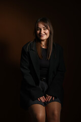 Beautiful young woman in stylish clothes sitting on chair against dark brown background