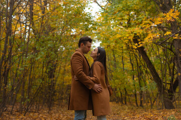 Romantic couple standing close together in autumn forest, wearing matching brown coats, looking into each other's eyes with love and tenderness surrounded by fall colors.
