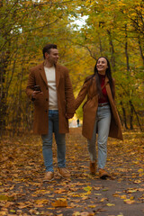 Happy couple walking hand in hand through autumn park, wearing matching brown coats and jeans, smiling and enjoying romantic fall day surrounded by colorful foliage.