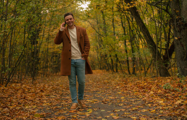Smiling man in brown coat walking through autumn forest, talking on smartphone, enjoying conversation outdoors among fallen yellow leaves and warm seasonal colors.