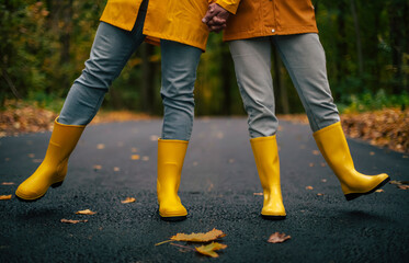 Playful couple holding hands and kicking feet in yellow rain boots on autumn forest road, wearing matching raincoats and jeans, enjoying fun and love outdoors in fall season.