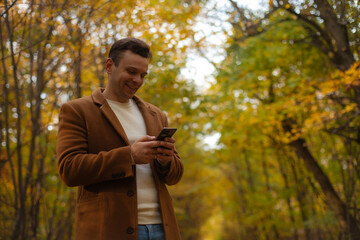 Smiling man in brown coat using smartphone in autumn park, standing among colorful trees and yellow leaves, enjoying online communication outdoors in fall season.