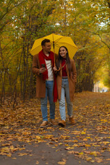 Happy couple walking together under a yellow umbrella in autumn park, smiling and talking, dressed in matching brown coats and jeans surrounded by colorful fall foliage.