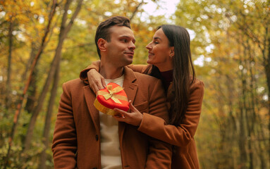 Romantic couple celebrating Valentine’s Day in an autumn forest. Woman gives a heart-shaped gift box to her partner, sharing a warm, loving moment together.