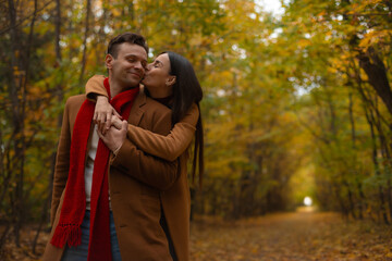 Loving couple in brown coats standing in autumn park, woman hugging and kissing man on cheek, both smiling warmly surrounded by golden fall trees and romantic atmosphere.