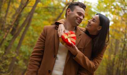 Romantic couple celebrating Valentine’s Day in an autumn forest. Woman hugs her partner and gives him a heart-shaped gift box, sharing a warm and loving moment.