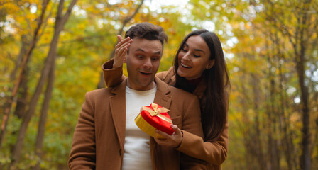 Happy couple celebrating Valentine’s Day in an autumn forest. Woman surprises her partner with a heart-shaped gift box, and he reacts with joy and excitement.
