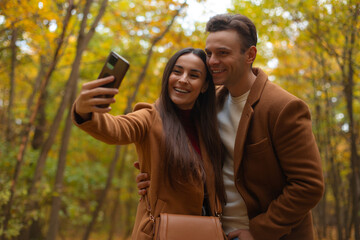 Happy couple taking a selfie in an autumn forest, smiling and posing closely together. Romantic fall atmosphere perfect for Valentine’s Day themes and love-related content.