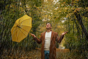 Smiling young man in brown coat standing in autumn rain with yellow umbrella in park, enjoying rainy weather and nature, expressing happiness and freedom outdoors.