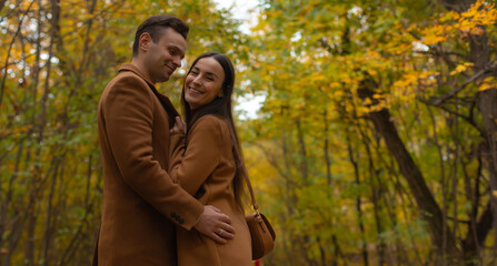 Romantic couple embracing in an autumn forest, smiling warmly at the camera. Cozy fall mood, love, affection, and Valentine’s Day atmosphere in a natural outdoor setting.