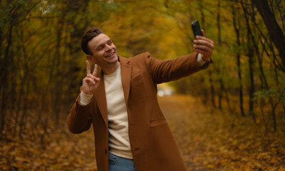 Smiling young man in brown coat taking selfie in autumn forest, showing peace sign with hand, enjoying outdoor walk among colorful fall foliage, cheerful lifestyle moment.