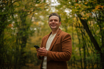 Smiling young man in brown coat holding smartphone in autumn forest, looking away with cheerful expression, surrounded by colorful fall foliage and natural light.