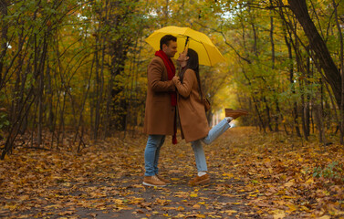 Romantic couple celebrating Valentine’s Day in an autumn forest, embracing under a yellow umbrella. Cozy fall mood, love, warmth, and happiness captured in a tender moment.
