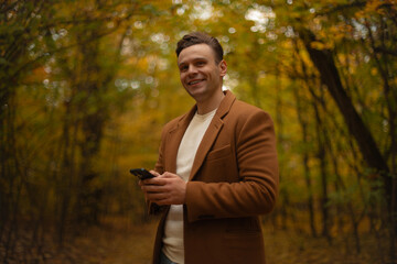 Smiling man in brown coat standing in autumn forest using smartphone, enjoying walk outdoors surrounded by colorful fall foliage and warm natural light.