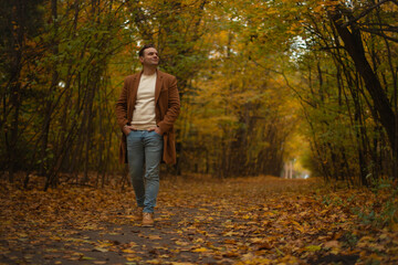 Confident man in brown coat and jeans walking through autumn forest path covered with golden leaves, enjoying peaceful fall day and nature outdoors.