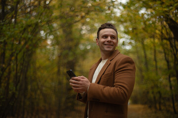 Smiling man in brown coat holding smartphone in autumn forest, enjoying a peaceful moment surrounded by golden foliage and soft natural light, symbolizing connection and calmness.