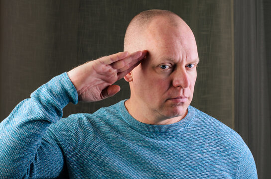 A bald man in a blue sweater is saluting with his right hand, looking directly at the viewer