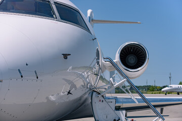Close-up of  a business jet showing turbofan engine, fuselage, cockpit windows, and deployed boarding stairs. Aircraft parked on tarmac with antenna visible. Clear blue sky.