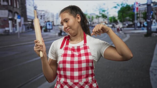 Woman cook holding a rolling pin and showing thumbs down gesture on a city street wearing red gingham apron and tshirt; kitchen disappointment.