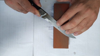 Closeup view of hands sharpening a metal knife on a brown whetstone, showing the technique and texture of the blade and sharpening surface.