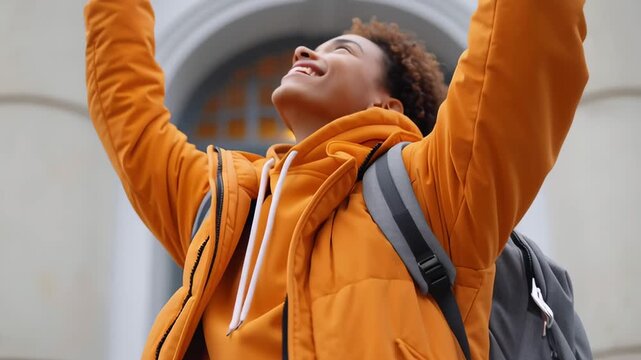 A young man in a bright orange jacket is smiling and holding his arms out wide. He is wearing a backpack and he is happy and excited