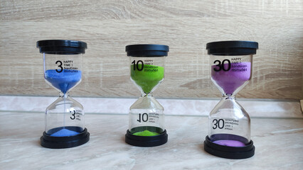 Three hourglasses with blue, green, and purple sand display 3, 10, and 30 minute timers, arranged neatly on a light surface with a wood-textured background.