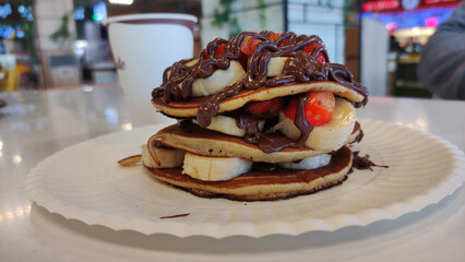 Close up of layered pancakes filled with bananas, strawberries, and thick chocolate drizzle, served on a paper plate in a cozy cafe setting.