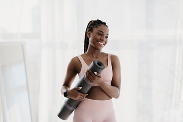 A young black woman smiles while holding a fitness mat in her home gym. She is dressed in athletic wear, ready for a yoga or pilates session. Bright light fills the indoor space.