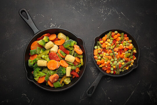 Preparing healthy meal with vibrant frozen mixed vegetables in cast iron pans on a dark background, top view